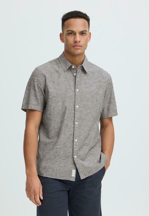 Homme aux cheveux courts et bouclés portant une chemise grise à manches courtes boutonnée et un pantalon foncé, debout avec une main dans la poche.