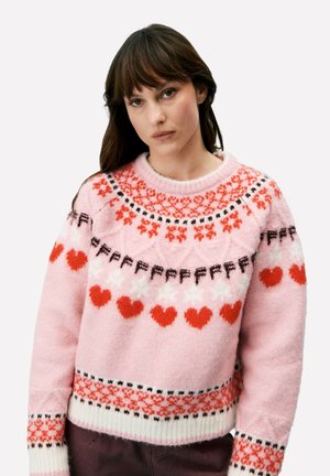 Vrouw die een roze gebreide trui draagt met rode harten en geometrische patronen, staand tegen een effen lichte achtergrond.