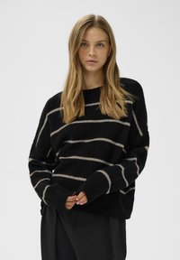 Suéter negro de punto con rayas horizontales beige, mangas amplias y un corte relajado. La textura suave y los puños acanalados mejoran la comodidad.