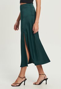 Grüne satin Maxi-Rock mit seitlichen Schlitzen, hohem Taillendesign und glatter Textur. Kombiniert mit schwarzen Riemchen-Heels und einem engen schwarzen Oberteil.