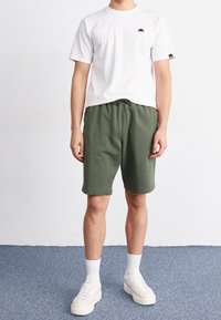 Hvid t-shirt med korte ærmer og et lille rødt logo, parret med grønne sweatshorts og hvide sneakers med pastelfarver.