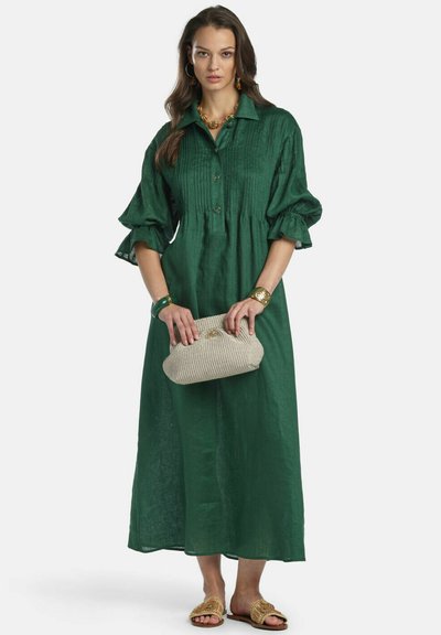 Mujer con un vestido largo verde abotonado con mangas abullonadas, que sostiene un bolso de mano beige tejido y lleva joyas doradas y sandalias.