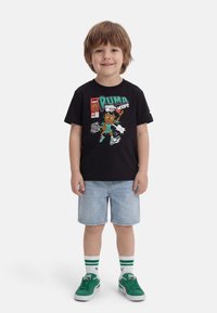 Νεαρός αγόρι όρθιος, φοράει μαύρο γραφικό μπλουζάκι με σχέδιο καρτούν μπάσκετ, ανοιχτόχρωμα τζιν σορτς, λευκές κάλτσες με πράσινες ρίγες και πράσινα παπούτσια.