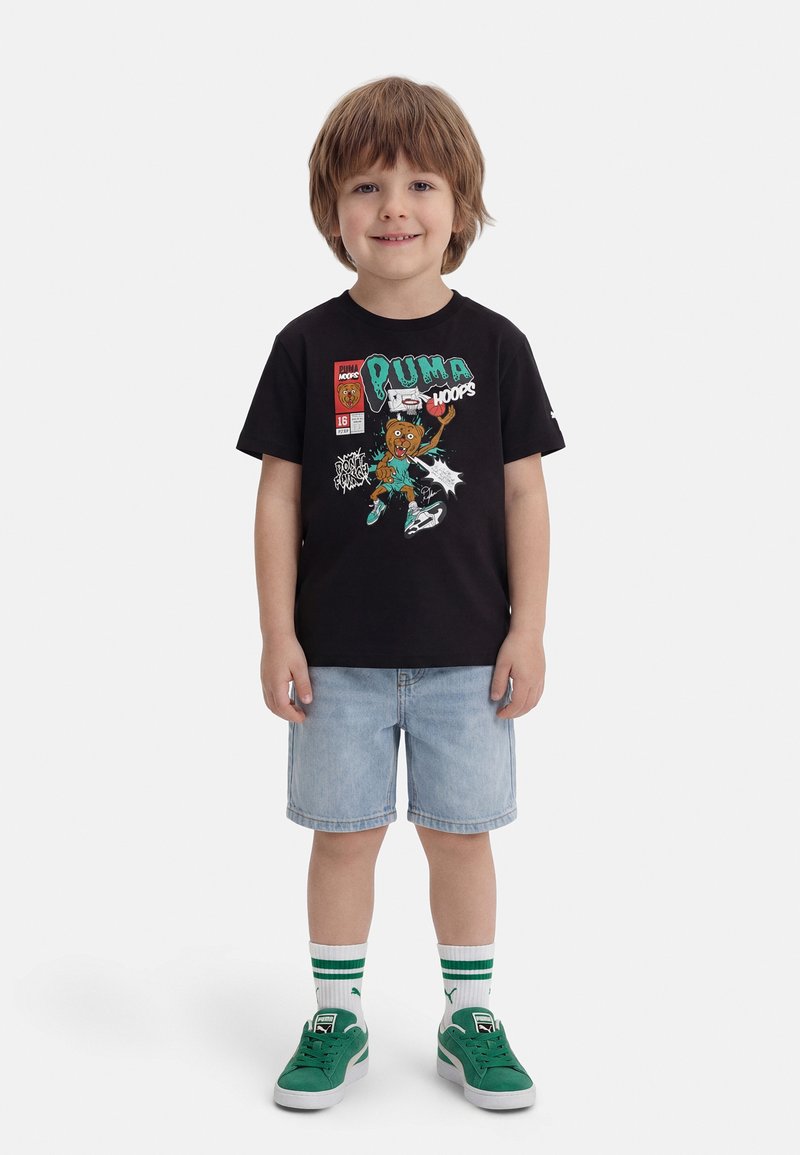 Νεαρός αγόρι όρθιος, φοράει μαύρο γραφικό μπλουζάκι με σχέδιο καρτούν μπάσκετ, ανοιχτόχρωμα τζιν σορτς, λευκές κάλτσες με πράσινες ρίγες και πράσινα παπούτσια.