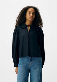 Blusa nera con colletto a scallop, fiocco e maniche puff testurizzate. Abbinata a jeans azzurri. Design semplice e pulito.