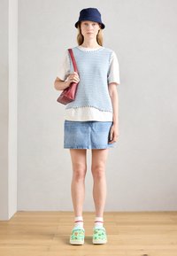 Himmelblaues, gestricktes, ärmelloses Oberteil über einem weißen T-Shirt, kombiniert mit einem Jeansrock. Accessoires sind eine rote Umhängetasche und grüne Clogs.