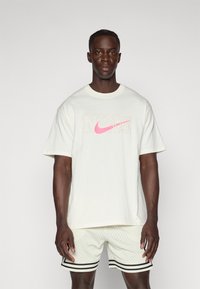 Bílá bavlněná tričko s velkým růžovým logem Nike a textem "NUKE" v světle béžovém písmu, doplněné o světlé šortky se černými pruhy.