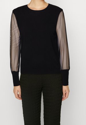 Black sweater with sheer, long sleeves and ribbed cuffs; round neckline. Textured dark green pants with a crinkled pattern.