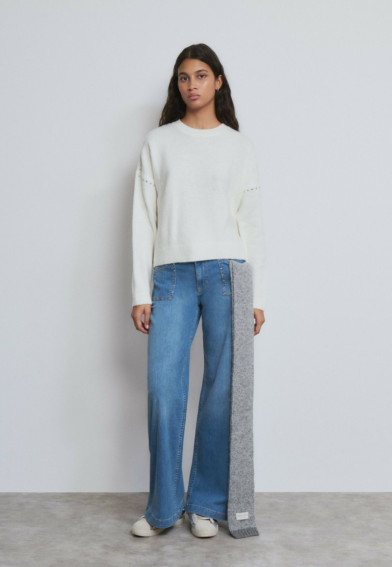 Frau mit langen dunklen Haaren, die einen weißen Pullover und weite blaue Jeans trägt und einen langen grauen Schal vor einfachem Hintergrund hält.