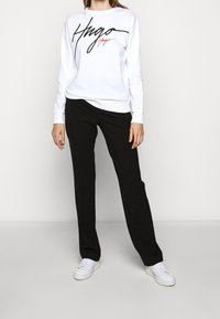 Felpa bianca con logo "Hugo" in nero e rosso, abbinata a pantaloni neri e sneakers bianche. Design semplice con una vestibilità rilassata.