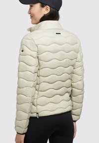 Jachetă pufoasă crem cu un model ondulat de quilt, guler înalt și nasturi de accent negri. Are un design elegant, ajustat și buzunare laterale.