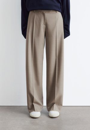 Beige wide-leg trousers worn with white shoes and a dark top, standing against a plain light wall and concrete floor.