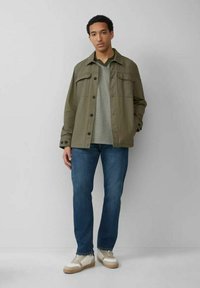 Olivgrüne Jacke mit Knopfverschluss und Fronttaschen, graues V-Ausschnitt-Shirt, blaue Jeans und beige Sneaker mit strukturierten Akzenten.