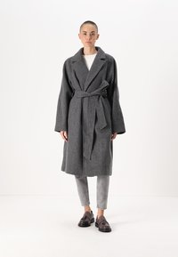 Siv dodatan plašč iz volne z pasom, velikimi reverji in dolgimi rokavi, v kombinaciji s svetlosivimi ozkimi kavbojkami in temnimi čevlji.