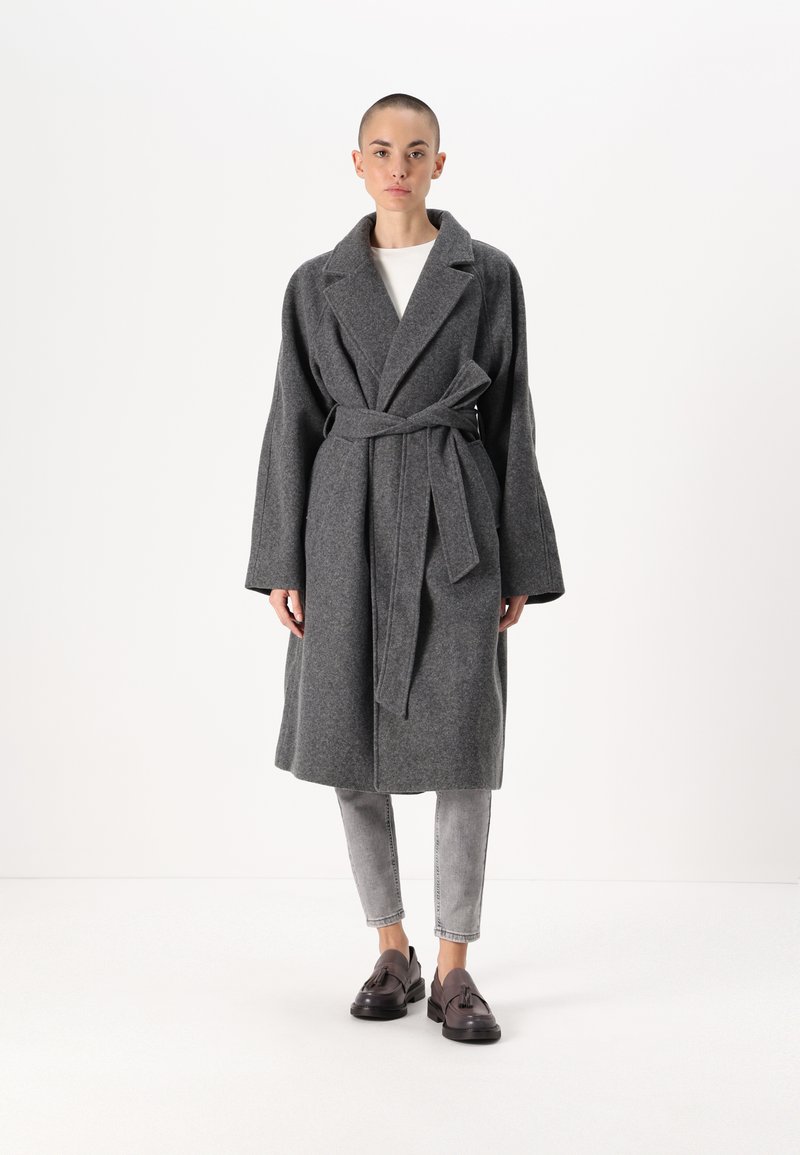 Siv dodatan plašč iz volne z pasom, velikimi reverji in dolgimi rokavi, v kombinaciji s svetlosivimi ozkimi kavbojkami in temnimi čevlji.