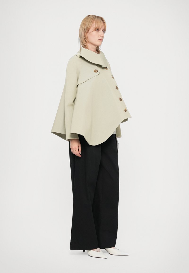 Veste cape vert clair avec un col surdimensionné et un devant à boutons proéminents, associée à un large pantalon noir et des chaussures blanches pointues.