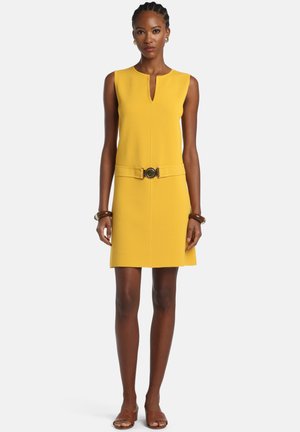 Woman wearing sleeveless yellow dress with front notch neckline, belted waist, brown bracelets, and brown open-toe sandals on white background.