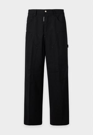Homme portant un manteau jaune moutarde, une chemise blanche, un pantalon large noir et des chaussures rouges, debout avec une main dans la poche sur un fond uni.