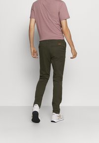 Olivinvihreät kapealahkeiset chinot, joissa on takataskun yksityiskohta, yhdistetty vaaleanpunaiseen T-paitaan ja valkoisiin lenkkareihin. Sileä rakenne.