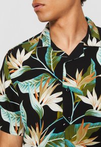 Personne portant une chemise noire à manches courtes avec un imprimé floral tropical coloré, se concentrant sur le col et le haut de la poitrine.