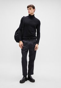 Suéter preto de gola alta, com um design ajustado, combinado com calças escuras e justas e botas pretas robustas. Modelo segura uma mala de mão texturizada.