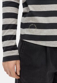 Langärmliges gestreiftes Hemd aus grauer und schwarzer Baumwolle, mit einem runden Logodetail am Saum und einem Rundhalsausschnitt. Kombiniert mit schwarzen Hosen.