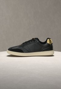 Melns sporta apavu pāris ar gludu ādas augšējo daļu, teksturētām zelta detaļām uz papēža un bēša gumijas zolī. Ietver tradicionālas auklas un izšuvumu detaļas.