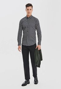 Koszula zapinana na guziki w kolorze granatowym z białym wzorem paisley, w połączeniu z czarnymi spodniami i błyszczącymi czarnymi butami, trzymająca zielony sweter.