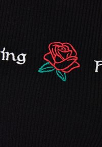 Svart ribbat tyg med röd rosbroderi och gröna blad, tillsammans med vit sytt text. Texten lyder: "Känsla." och "Fin."