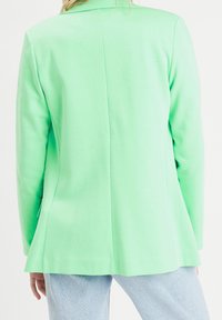 Blazer vert en tissu lisse, avec une coupe cintrée, une fente unique à l'arrière et des revers élégants, porté avec un jean bleu clair.