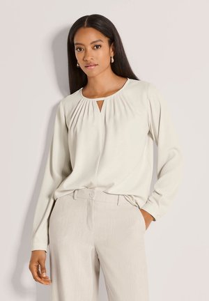Frau trägt eine cremefarbene Bluse mit langen Ärmeln und Schlüssellochausschnitt sowie hellbeige Hose, steht vor einem schlichten, hellen Hintergrund.