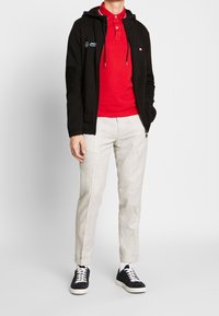 Uomo che indossa una felpa nera con zip, una polo rossa, pantaloni beige chiaro, calzini bianchi e sneakers nere, in piedi contro uno sfondo semplice.