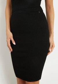 Jupe crayon noire côtelée en tissu extensible, avec un design ajusté et un logo accentué à la taille. Texture lisse et silhouette élégante.