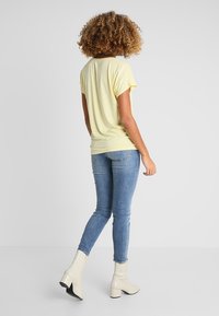 T-shirt giallo a maniche corte abbinato a jeans skinny blu chiaro e stivaletti beige alla caviglia; modella di spalle, in evidenza i dettagli dell'outfit casual.