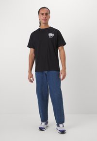 Tricou din bumbac negru cu mâneci scurte și gât rotund, având un imprimeu grafic alb pe partea stângă a pieptului. Purtați cu blugi din denim albastru.
