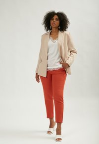 Beige blazer over een wit topje met kanten rand, gecombineerd met oranje slim-fit broek en witte open teen hakken. Gladde stof en op maat gemaakte pasvorm.