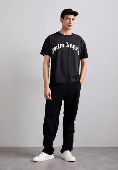 Black t-shirt with white "Palm Angels" logo, paired with black sweatpants featuring white side stripes and white sneakers.