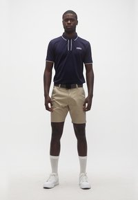 Άνδρας που φοράει μπλε μπλουζάκι πόλο, μπεζ σορτς με ζώνη, λευκές κάλτσες και λευκά αθλητικά παπούτσια, στέκεται μπροστά σε ένα απλό ανοιχτό φόντο.