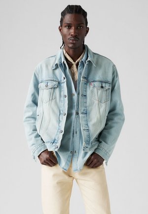 Light blue denim jacket with a button-up front, two chest pockets, and stitched seams, layered over a light shirt and cream-colored pants.