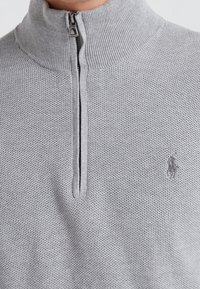 Grå zip-sweater med hög krage, texturerad stickad mönster och subtilt broderad logotyp på vänster sida. Dragkedjan har en metallisk finish.