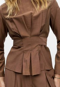 Brown, pleated top with a fitted waist and long sleeves, made of textured fabric. Features subtle stitching and a structured design.