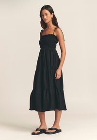 Abito lungo nero a balze con corpetto arricciato, spalline sottili e tessuto strutturato. Indossato con sandali neri piatti.