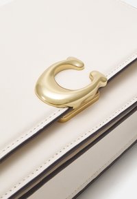 Borsa in pelle color crema con design strutturato, caratterizzata da una fibbia in metallo dorato prominente a forma di "C" e dettagli neri a contrasto.