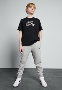 Černé tričko Nike s logem a kovovým potiskem "SB", doplněné šedými tepláky s malým logem a bílými teniskami s černými akcenty.