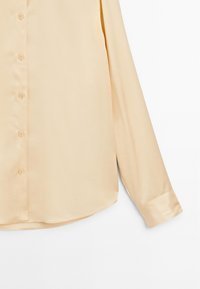 Chemise beige clair à boutons avec une finition satinée lisse, dotée d'un col pointu, de manches longues et d'un ourlet arrondi.