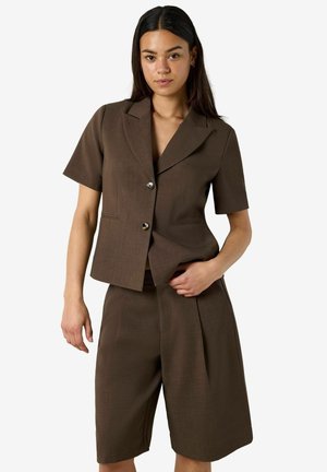 Jeune femme portant un blazer marron à manches courtes avec deux boutons et un short large assorti longueur genou sur fond uni.