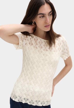 Kvinde med brunt hår iført guldringeøreringe og en kortærmet beige blomstret blondebluse, poserende med den ene hånd på halsen.