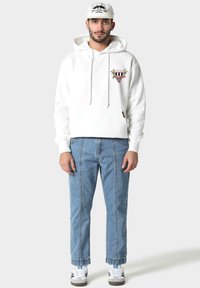 Uomo che indossa un felpa bianca con grafica, jeans azzurri chiari con cuciture verticali, cappellino bianco con emblema nero e sneakers bianche con strisce nere.