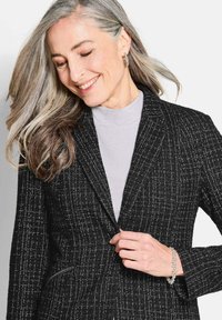 Schwarzer und weißer strukturierter Blazer mit Reverskragen, Knopfverschluss und seitlichen Reißverschlusstaschen, kombiniert mit einem hellgrauen Oberteil.