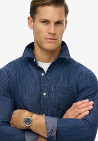 Denim button-up shirt met een spreidkraag, blauwe kleur, borstzak; gecompleteerd met een goudkleurig horloge op een bruine leren band.